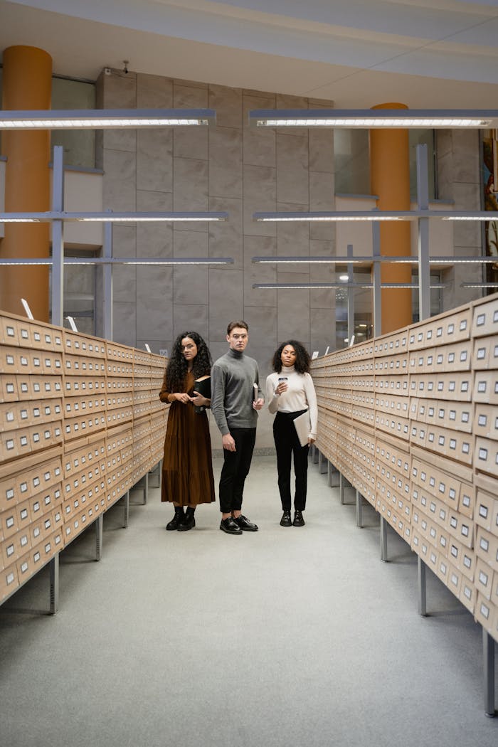 Three students browsing a card catalog aisle in a modern library setting.